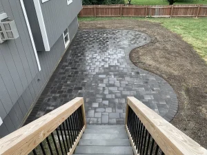 Curved dark paver patio extends from house foundation and stairs, surrounded by prepped soil and lawn, ideal for custom backyard entertainment space.