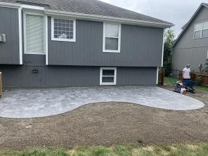Freshly installed light gray patio with a flowing edge layout sits behind a home, ready for landscaping with soil and equipment still on site.