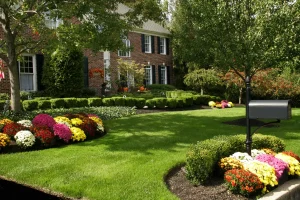 Front yard features vibrant flower beds, trimmed boxwoods, and a healthy green lawn in front of a red brick house with black shutters and white trim.