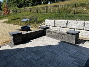 Paver patio features a built-in L-shaped seating wall with dark stone caps, set beside a white block retaining wall and a grassy slope with patio furniture.