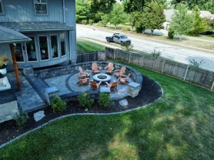 Circular fire pit area with Adirondack chairs on stone paver patio in fenced backyard near a gray house and residential street.