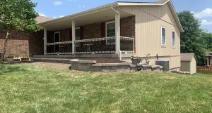 Raised stone flower bed with dark mulch along a covered porch of a beige and brick house on a sloped grassy lawn.